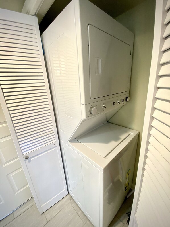 A compact laundry room with a stacked washer and dryer, a door with horizontal louvered panels, and a tiled floor.