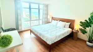 A bedroom with a large bed, a bedside table with a lamp, a potted plant, and a view of the sea through the window.
