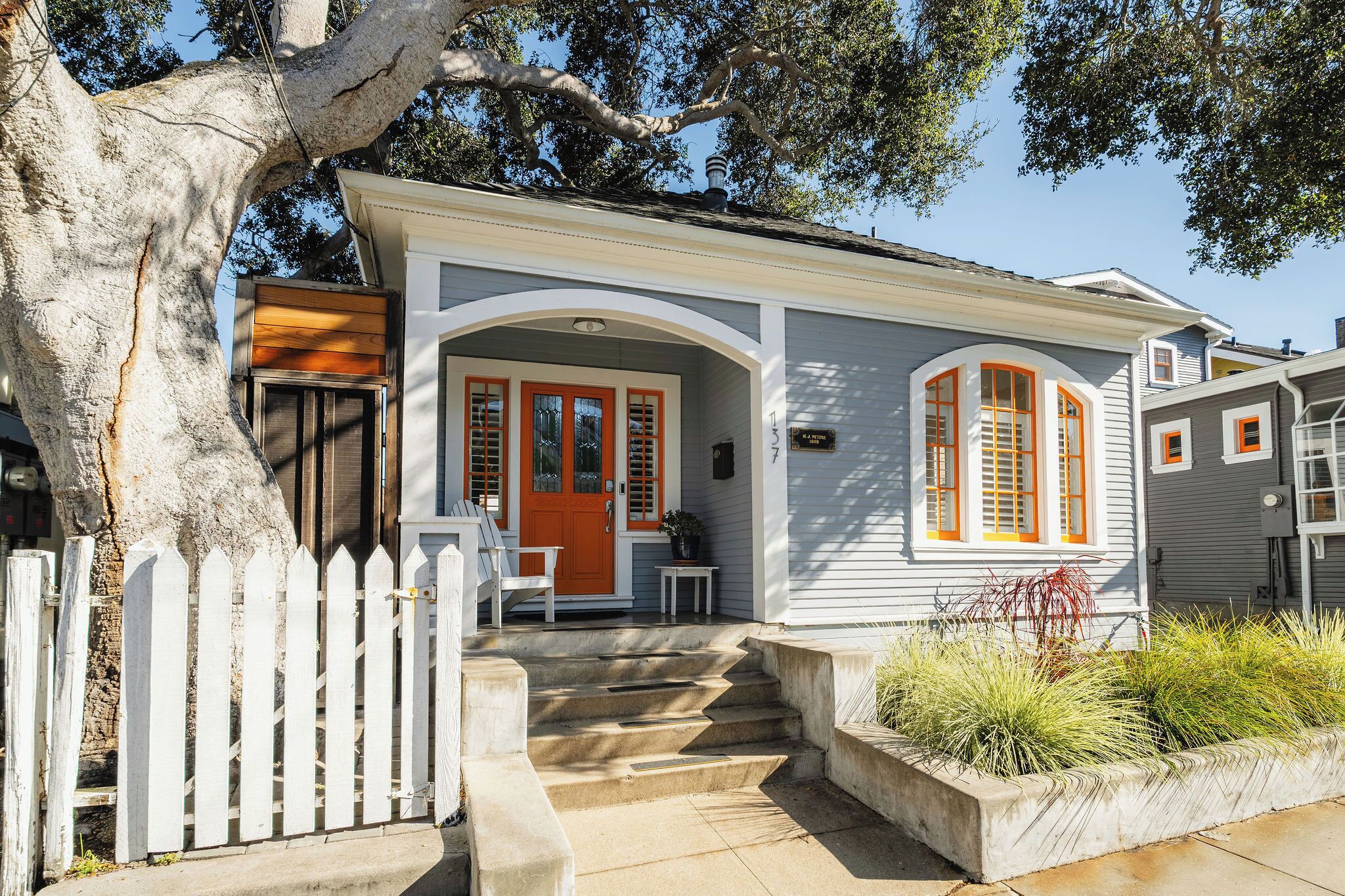 Une maison à deux étages avec une clôture blanche en bois, un porche à l’avant et un grand arbre.