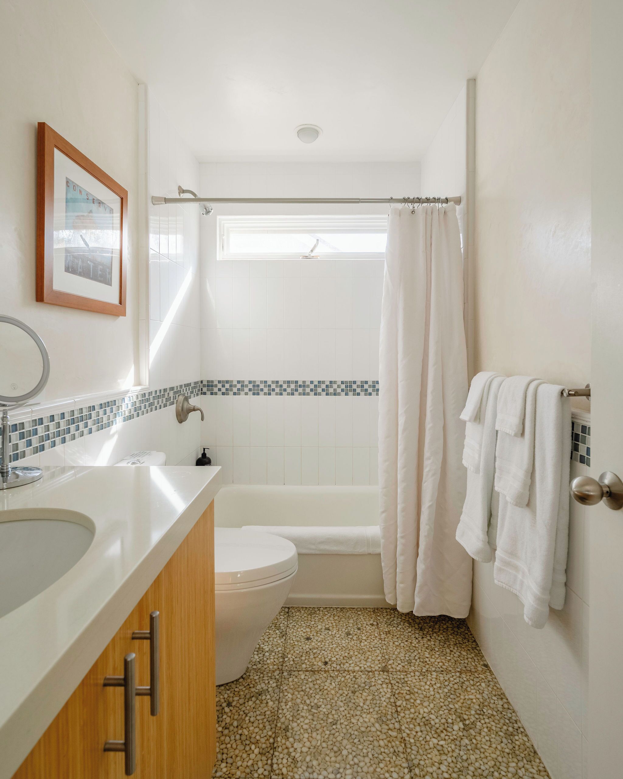 Ein Badezimmer mit einem weißen Waschbecken, einer Toilette und einer Badewanne. An der Wand hängt ein gerahmtes Bild und über der Badewanne befindet sich ein Fenster.
