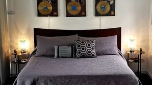Una cama bien hecha con una colcha oscura estampada, dos almohadas y un banco a juego. Encima de la cama, hay tres objetos decorativos fijados a la pared.