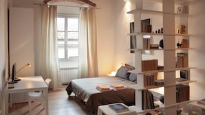Un dormitorio con cama, escritorio, silla, estantería y ventana con cortinas.