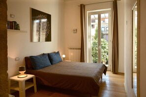 Ein Schlafzimmer mit einem Bett, einem Nachttisch mit Lampe, einem Wandbild und Blick durch eine Schiebetür auf einen Außenbereich.