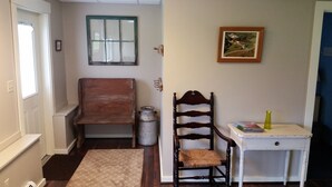 Un couloir avec un banc en bois, une chaise, une petite table avec des livres, un tableau accroché au mur et une fenêtre.