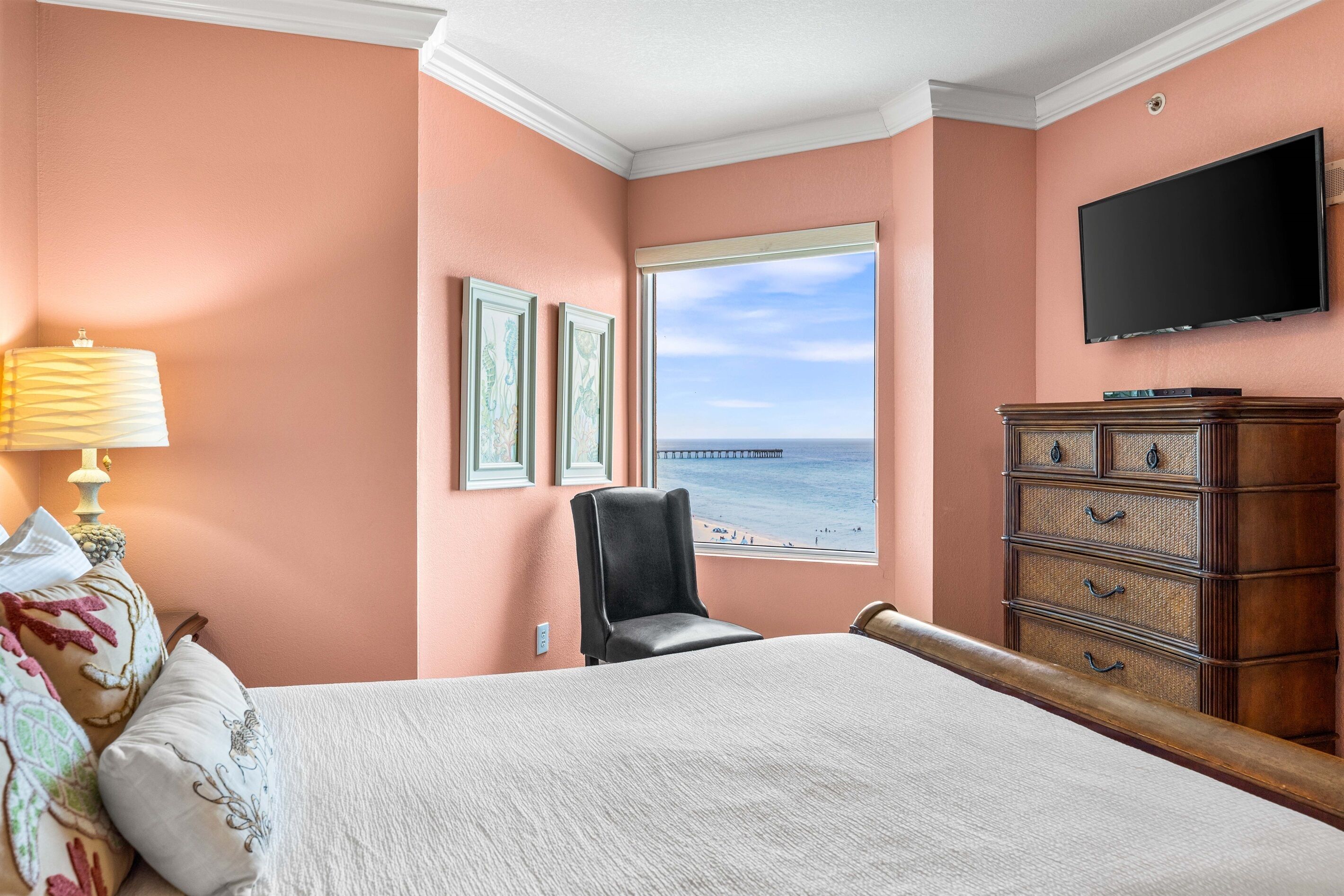 A bedroom with a bed, a chair, a dresser, a TV, and a window with a view of the beach.