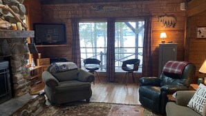 L’intérieur de ce chalet accueillant est doté d’une cheminée en pierre, d’un plafond en bois et d’une grande fenêtre donnant sur une forêt. On y trouve deux canapés, une table basse et un téléviseur fixé au mur.