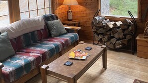 L’intérieur chaleureux d’un chalet avec un canapé en bois, une table avec des livres et une lampe, et une vue sur la forêt à travers de grandes fenêtres.