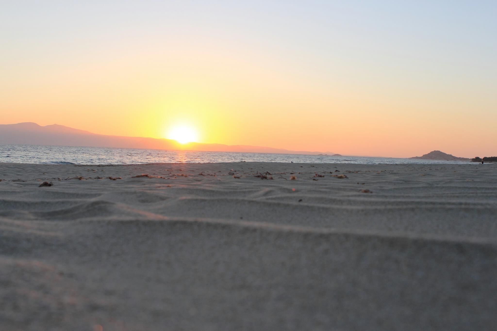 A beach with a sunset over the ocean, distant mountains, and scattered debris on the sand.