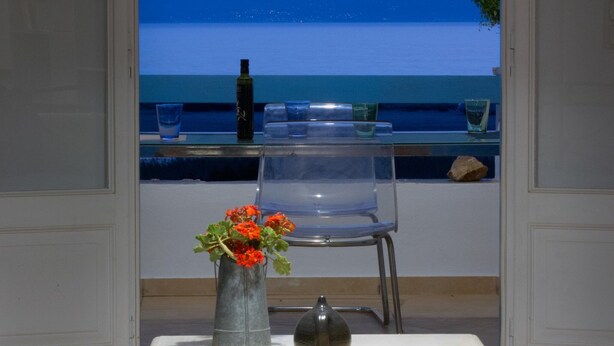A table with a vase of flowers, a bottle, and a chair by a window overlooking a body of water. - Breathtaking sunset, salt lake and Aegean sea view. 150m from magnificent beach. (NAXOS)