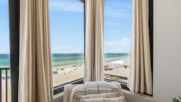 A room with a view of a beach, featuring a window with curtains, a bed with a blanket, and a pillow.