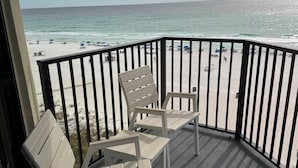 Ein Balkon mit zwei Holzstühlen und einem Tisch mit Blick auf einen Strand mit Sonnenschirmen und Menschen.