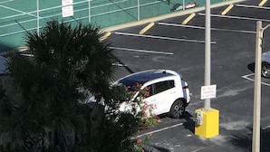 Ein Tennisplatz mit Zaun, ein kleines Gebäude und ein Parkplatz mit einem weißen Auto.