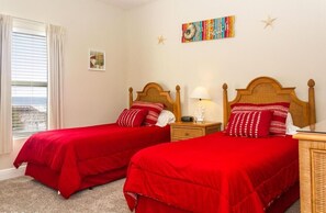 A room with two single beds, each with a red bedspread and matching pillows. There is a small bedside table with a lamp and a decorative item on each bed. The room has a window with white curtains, a framed picture on the wall, and a colorful artwork above the beds. - The Perfect Vacation Paradise With A Golf Front View (Fort Walton Beach)