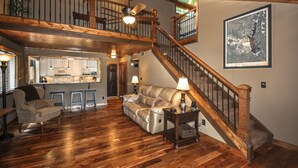 A spacious living area with a wooden staircase, a kitchen in the background, and a large framed artwork on the wall. - Lake Front, PETS, Theater, Dock, Swim-Fish-Boat,  Fire Pit, NEW BOAT, Pickleball (Waleska)