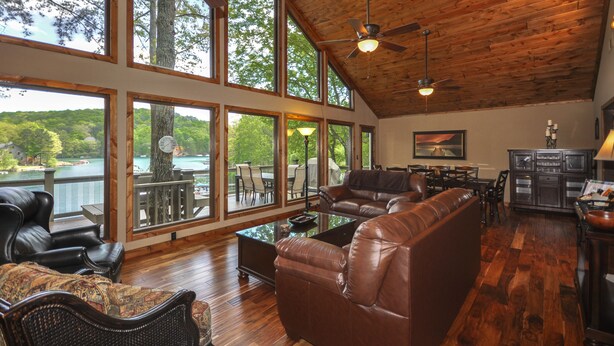 A spacious living room with leather sofas, a glass coffee table, and a dining area with chairs. Large windows offer views of a lake and trees. - Lake Front, PETS, Theater, Dock, Swim-Fish-Boat,  Fire Pit, NEW BOAT, Pickleball (Waleska)