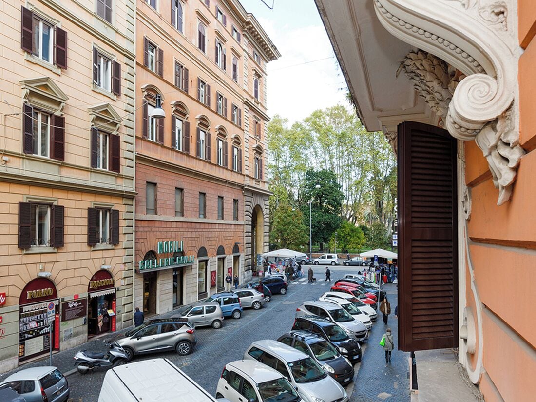 A street view with parked cars, a row of shops, and a building with a classical facade.