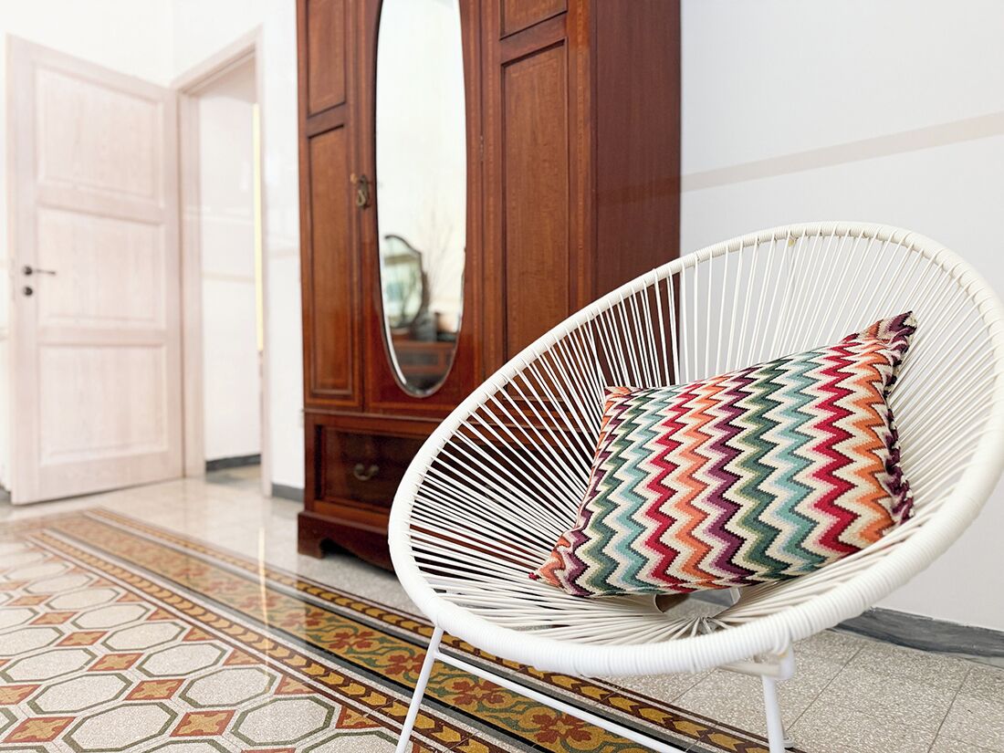 A white wicker chair with a colorful zigzag pillow in a room with a wooden cabinet and a large mirror.