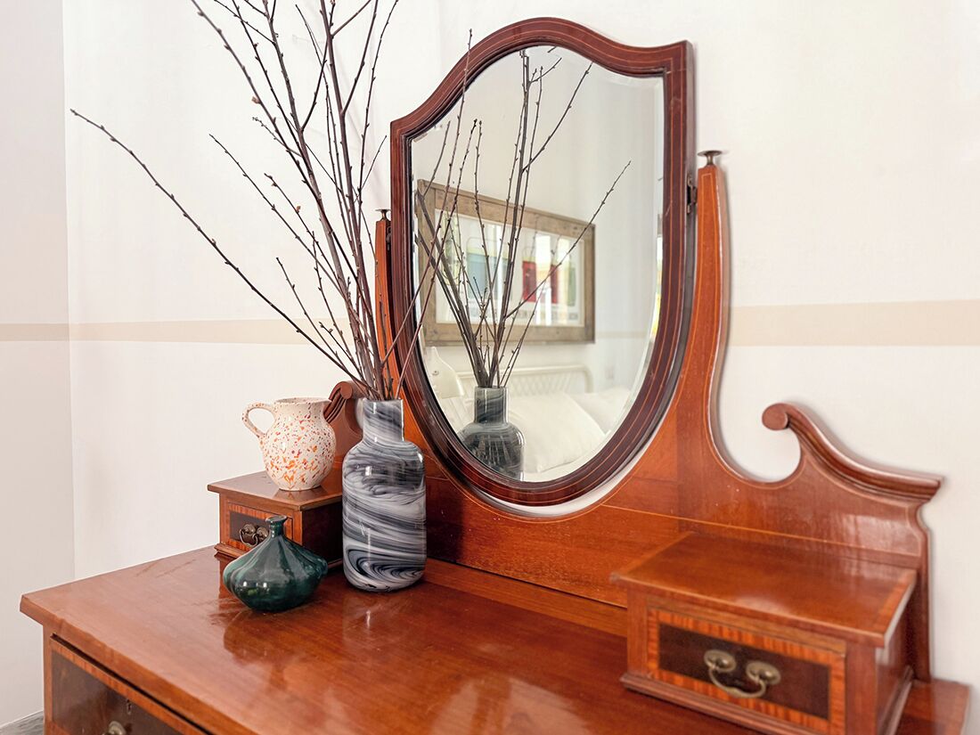 Een houten dressoir met een spiegel, een vaas met takken, en een keramieken kan.