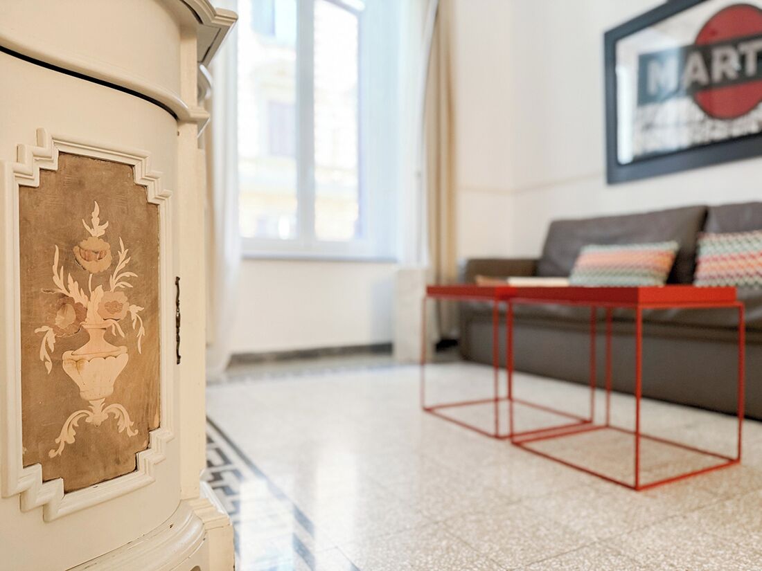 Een kamer met een witte muur, een gekaderd kunstwerk, een rode tafel en een grijze bank met gestreepte kussens.