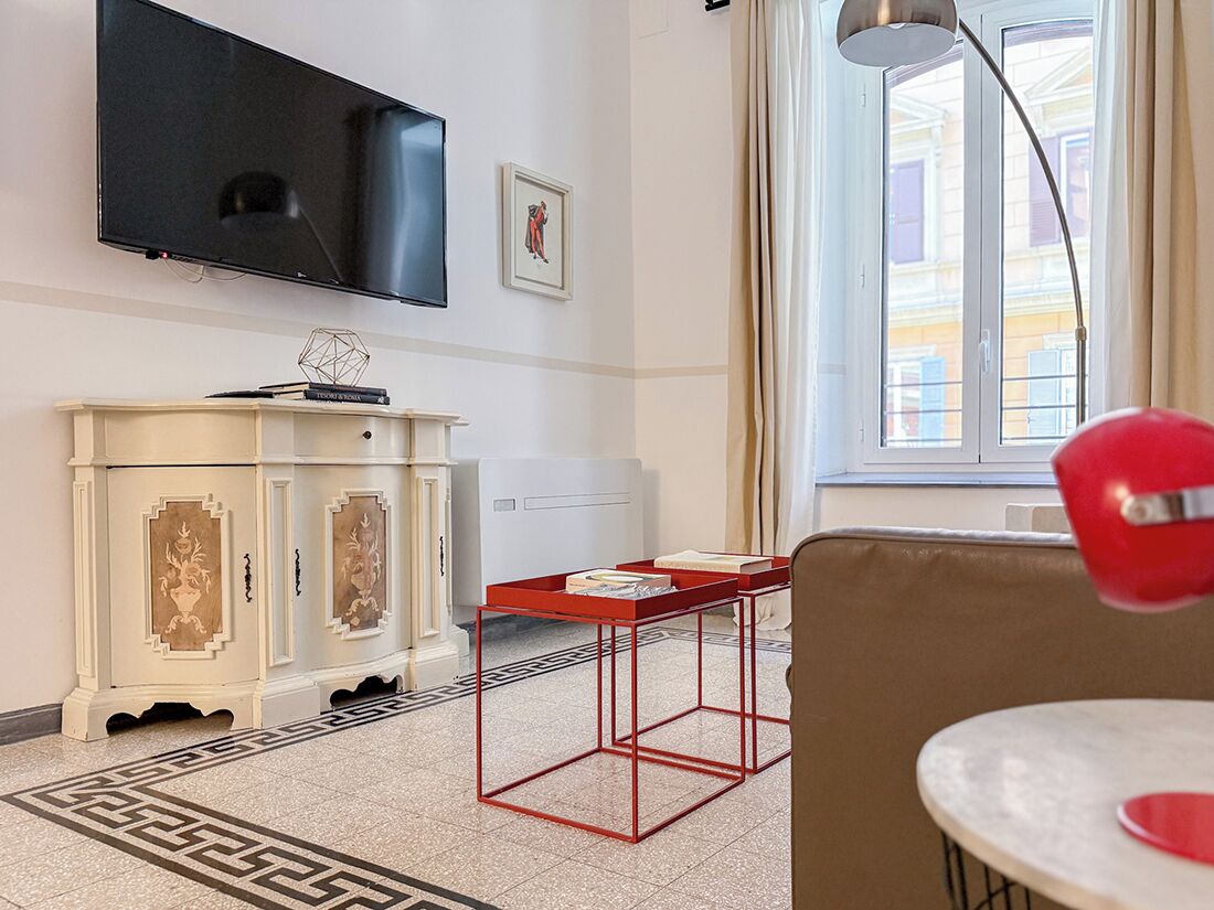 Une chambre d’hôtel moderne dotée d’un téléviseur à écran plat, d’une armoire blanche agrémentée de panneaux décoratifs, d’une table en métal rouge et offrant une vue sur la ville grâce à une grande fenêtre.