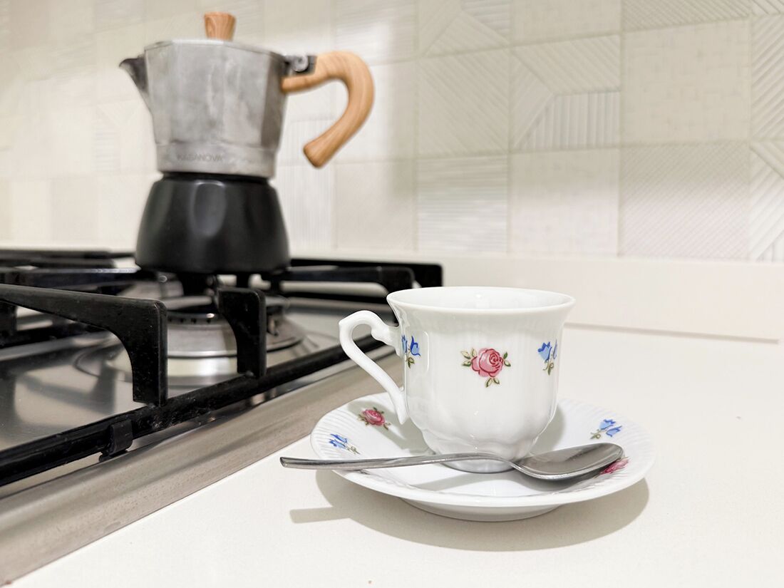 A white ceramic cup with floral designs on a matching saucer and spoon, placed on a kitchen counter with a gas stove and a coffee pot in the background.