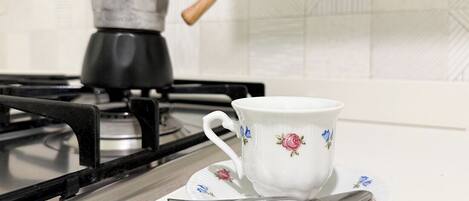 A white ceramic cup with floral designs on a matching saucer and spoon, placed on a kitchen counter with a gas stove and a coffee pot in the background.