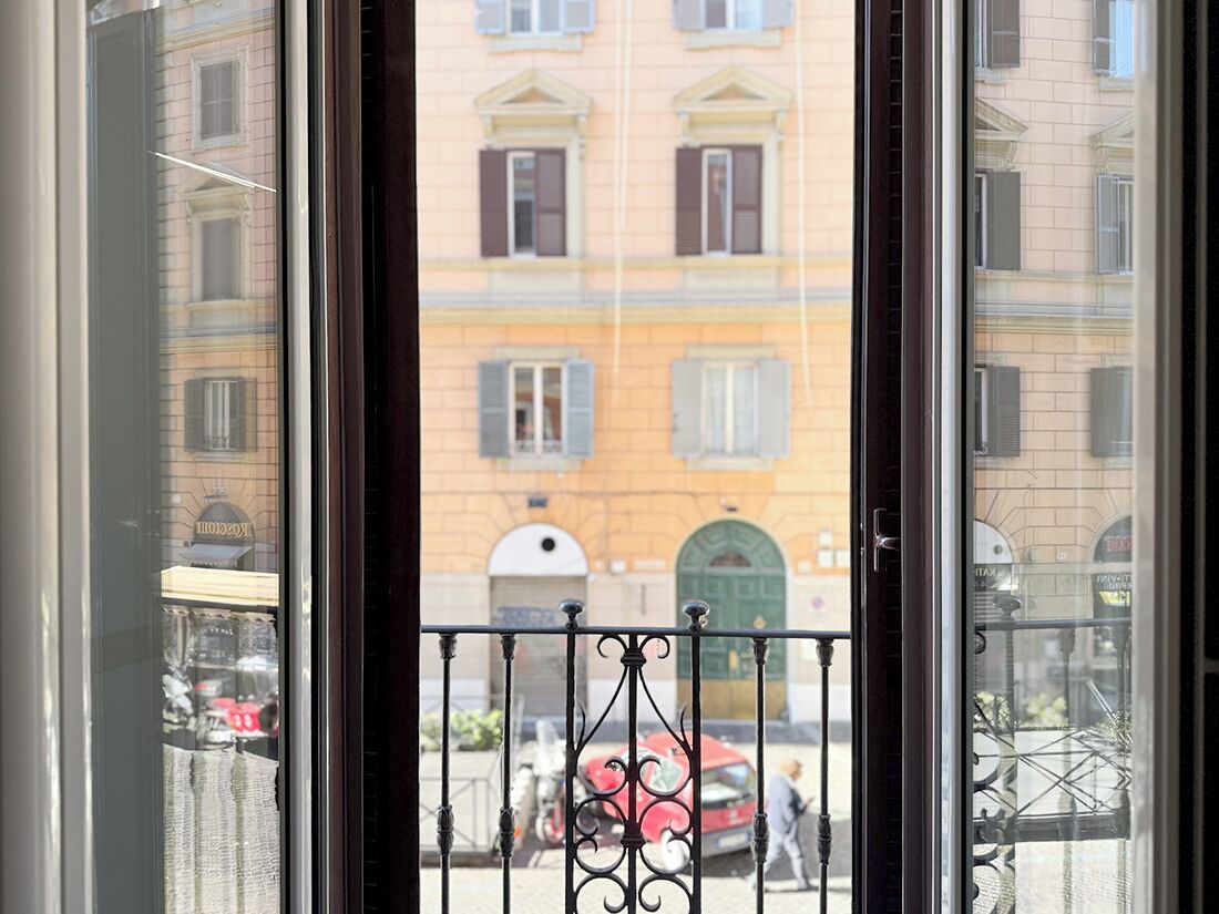 Une vue à travers une fenêtre ornée d’une rambarde en métal, une voiture rouge garée à l’extérieur, et un bâtiment doté de nombreuses fenêtres et volets.