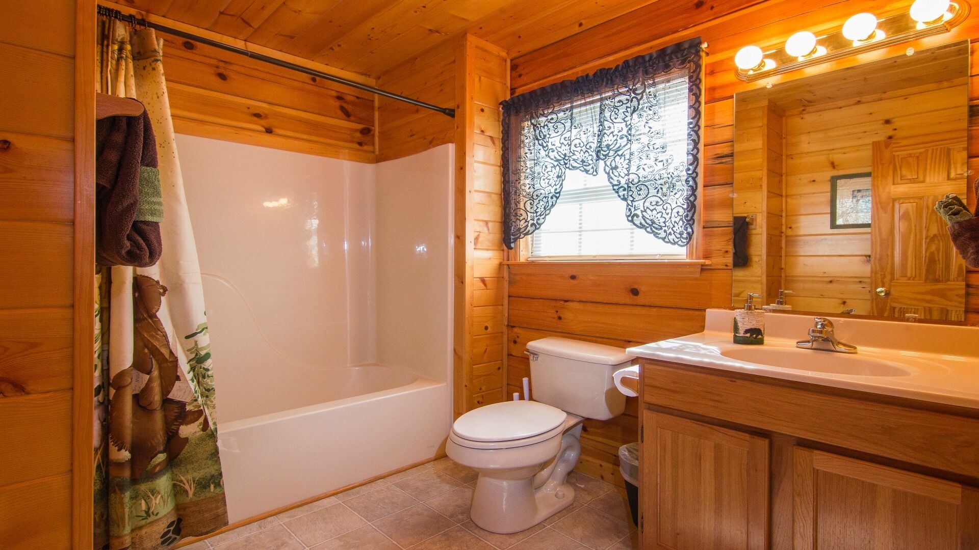 Une salle de bain rustique, aménagée dans une cabane en bois, avec une baignoire, des toilettes, un lavabo et une fenêtre ornée de rideaux en dentelle.