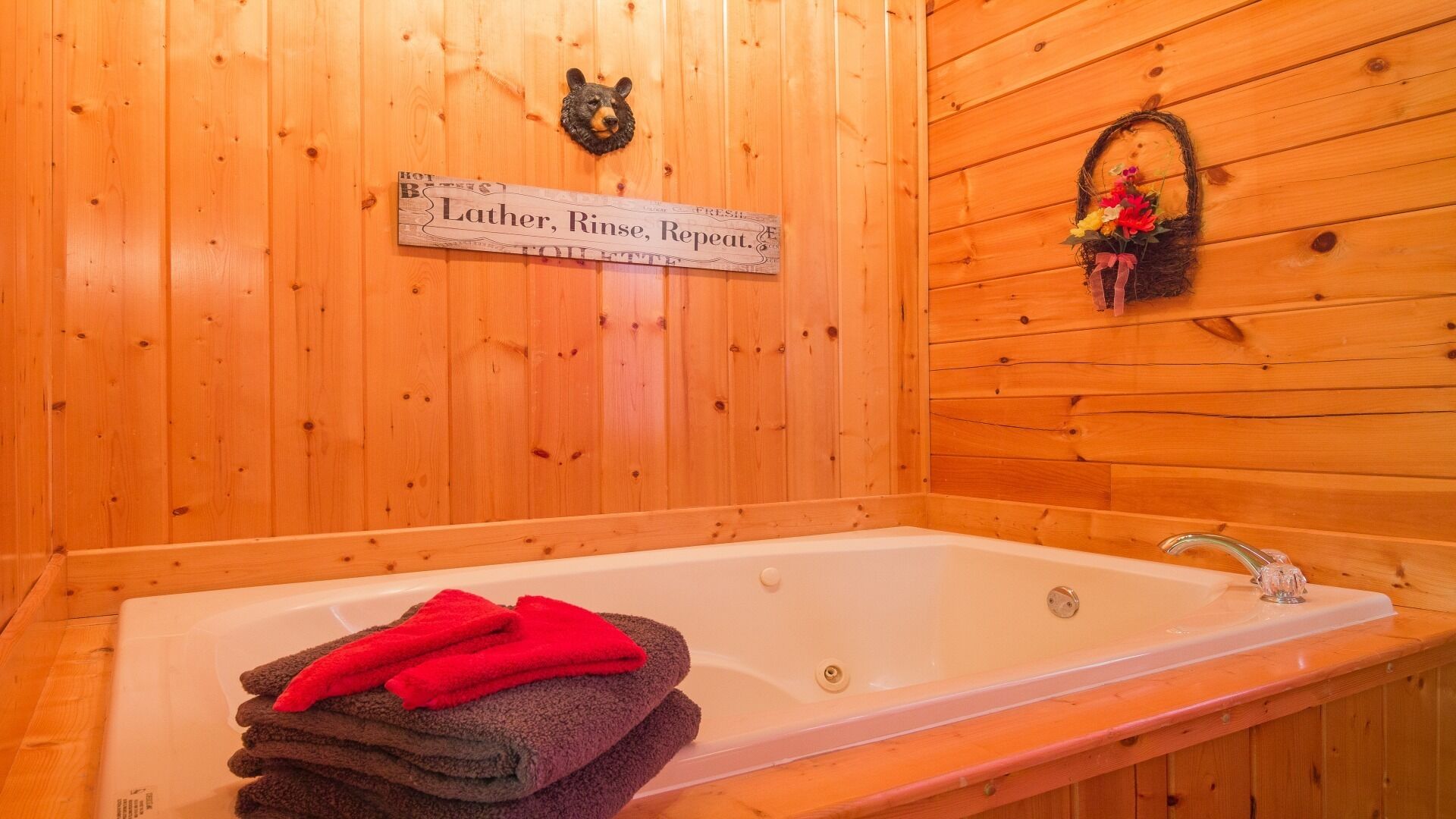 A wooden cabin bathroom with a bathtub, a bear head decoration, a sign reading "Lather, Rinse, Repeat.", and a basket with flowers.