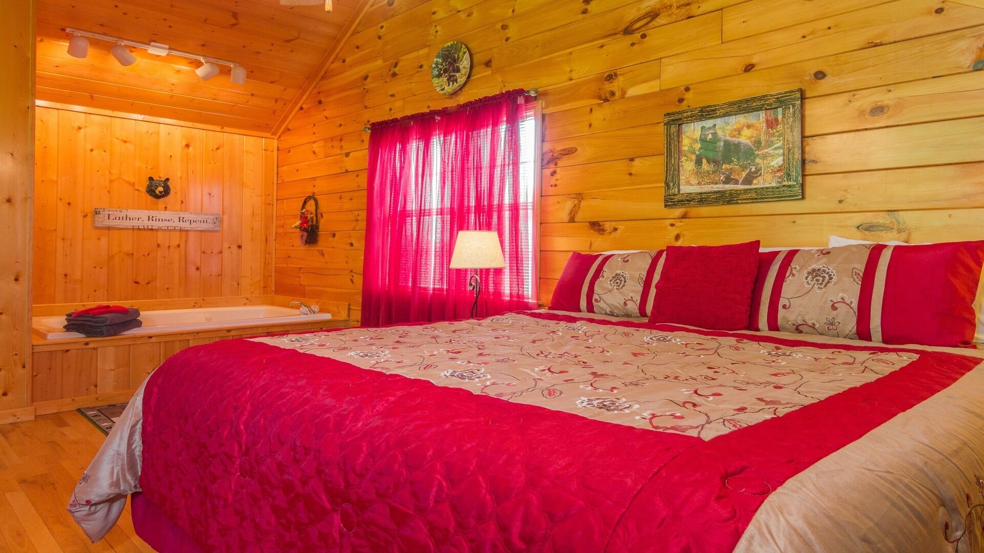 Habitación tipo cabaña de madera con una cama grande, una manta roja, un cartel que dice 'Lather, Rinse, Repeat', una pared de madera y una ventana con cortinas rojas.