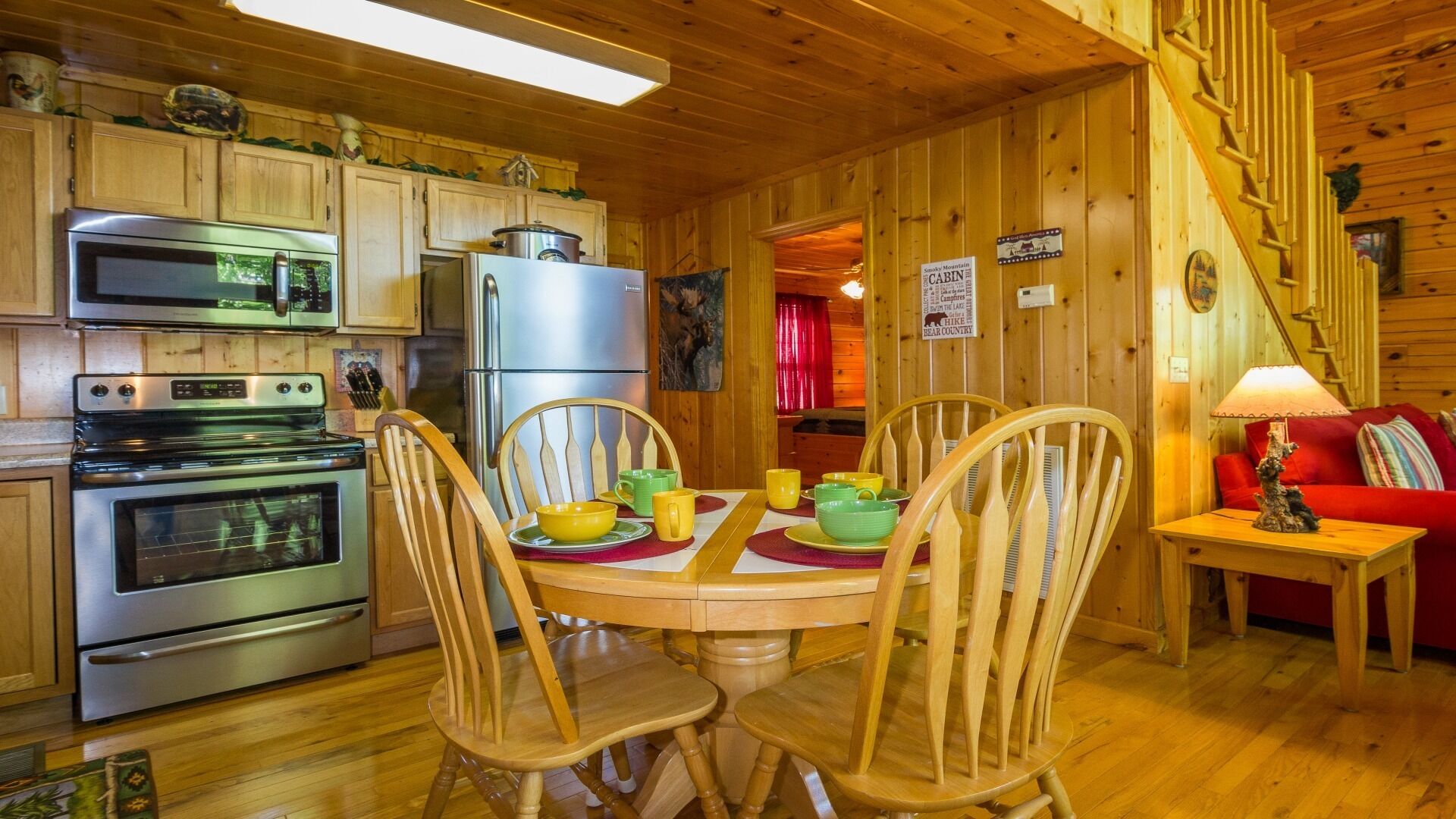 A cabin interior with wooden walls, a dining table with chairs, a kitchen with appliances, and a red sofa.
