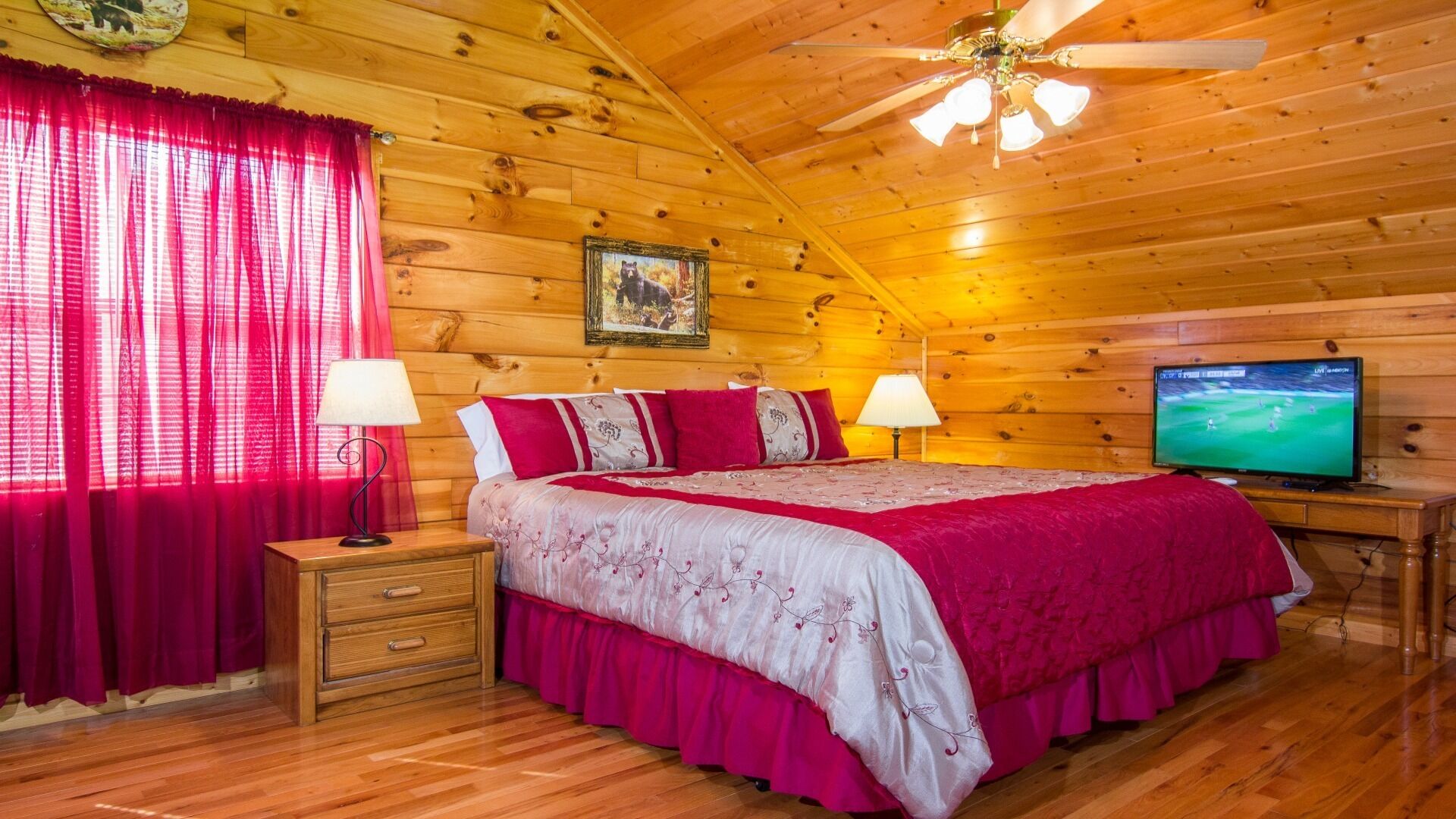 A cozy log cabin bedroom with a bed, two bedside tables, a ceiling fan, a television, and a painting on the wall.