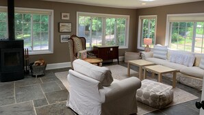 Un salon avec une cheminée, de grandes fenêtres, un canapé, une table basse et un pouf à motifs.