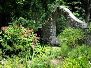Un jardín exterior con un arco de piedra rodeado de abundante vegetación y plantas en flor.