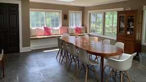 Un coin repas avec une grande table en bois, des chaises blanches et un banc intégré agrémenté de coussins roses.
