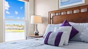 A neatly made bed with a white and purple pillow set, a bedside lamp, and a view of a residential area through a large window.