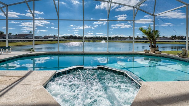A swimming pool with a hot tub, surrounded by a covered area with a view of a lake and houses in the background.