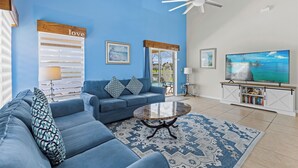 A living room with a blue wall, a ceiling fan, a sofa set, a coffee table, a TV, and a bookshelf.