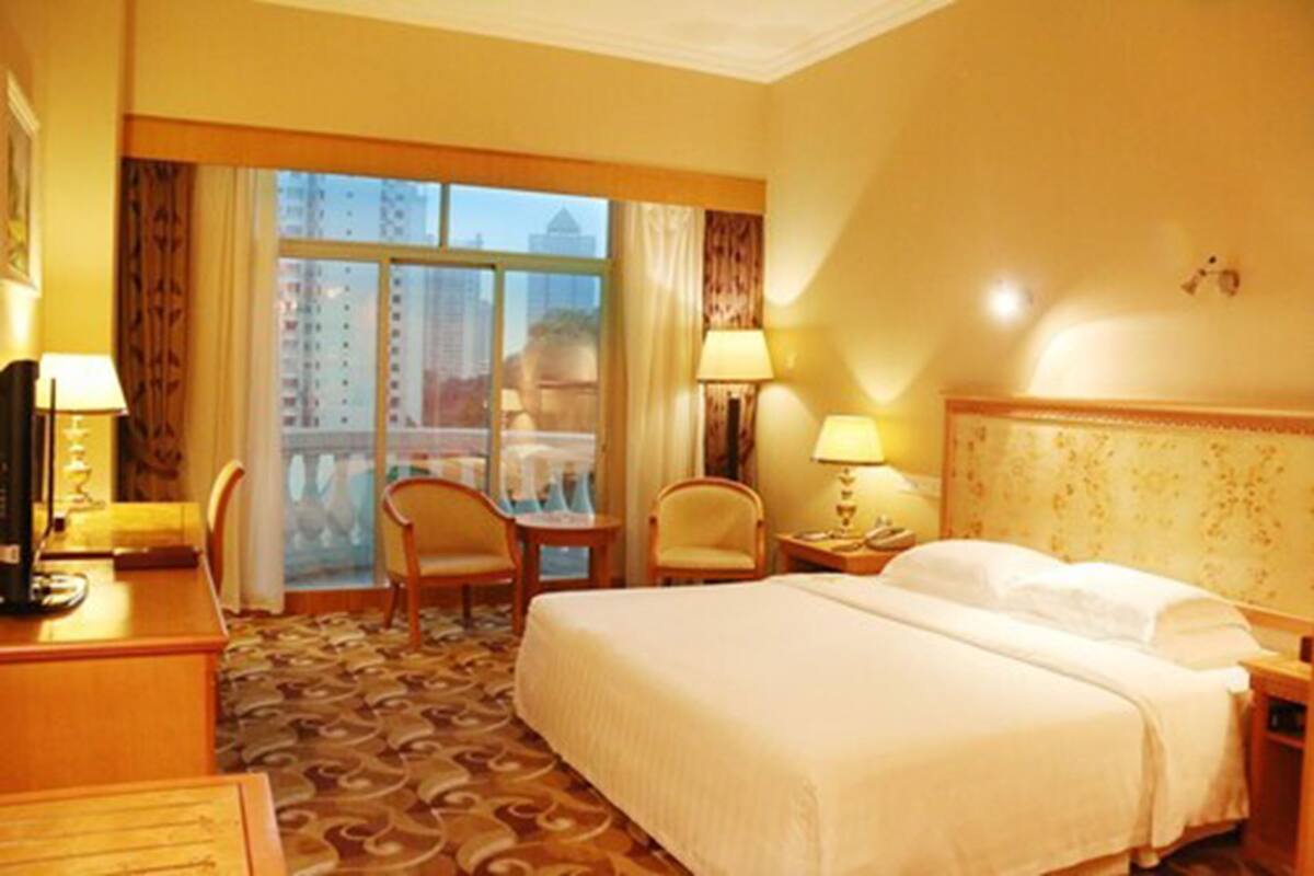 A hotel room with a bed, two chairs, a desk, a lamp, and a view of a cityscape through large windows.