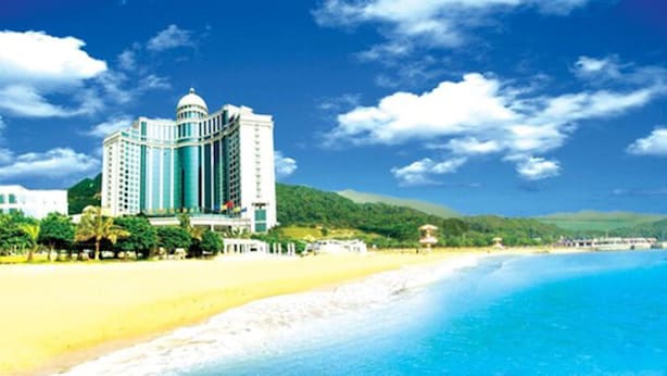 Een groot hotel met een centrale koepel en meerdere torens, gelegen aan een strand met helderblauw water en groene heuvels op de achtergrond.