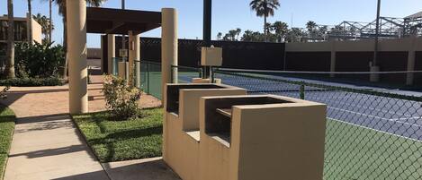 Una cancha de tenis con una red, rodeada por una valla y palmeras.