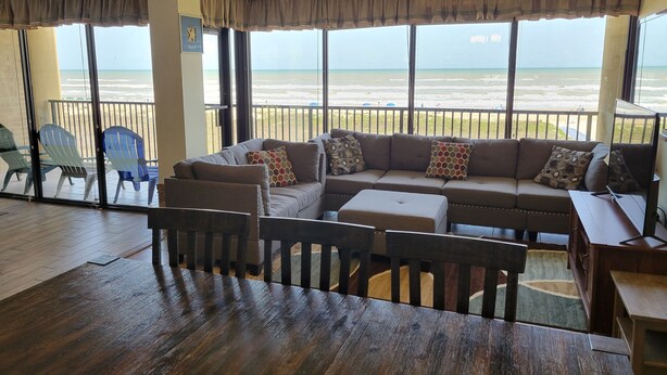 Amplia sala de estar con un sofá grande, una mesa de centro de madera y un televisor. Hay varias sillas y una mesita. La habitación tiene ventanas amplias con vista a la playa.