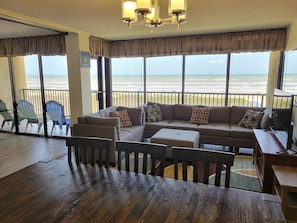 A spacious living area with a large sofa, a wooden coffee table, and a television. There are several chairs and a small table. The room has large windows with a view of the beach.