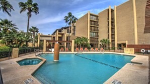 Zona de piscina de hotel con una piscina cristalina, sillas de descanso, palmeras y un edificio de varios pisos con balcones.
