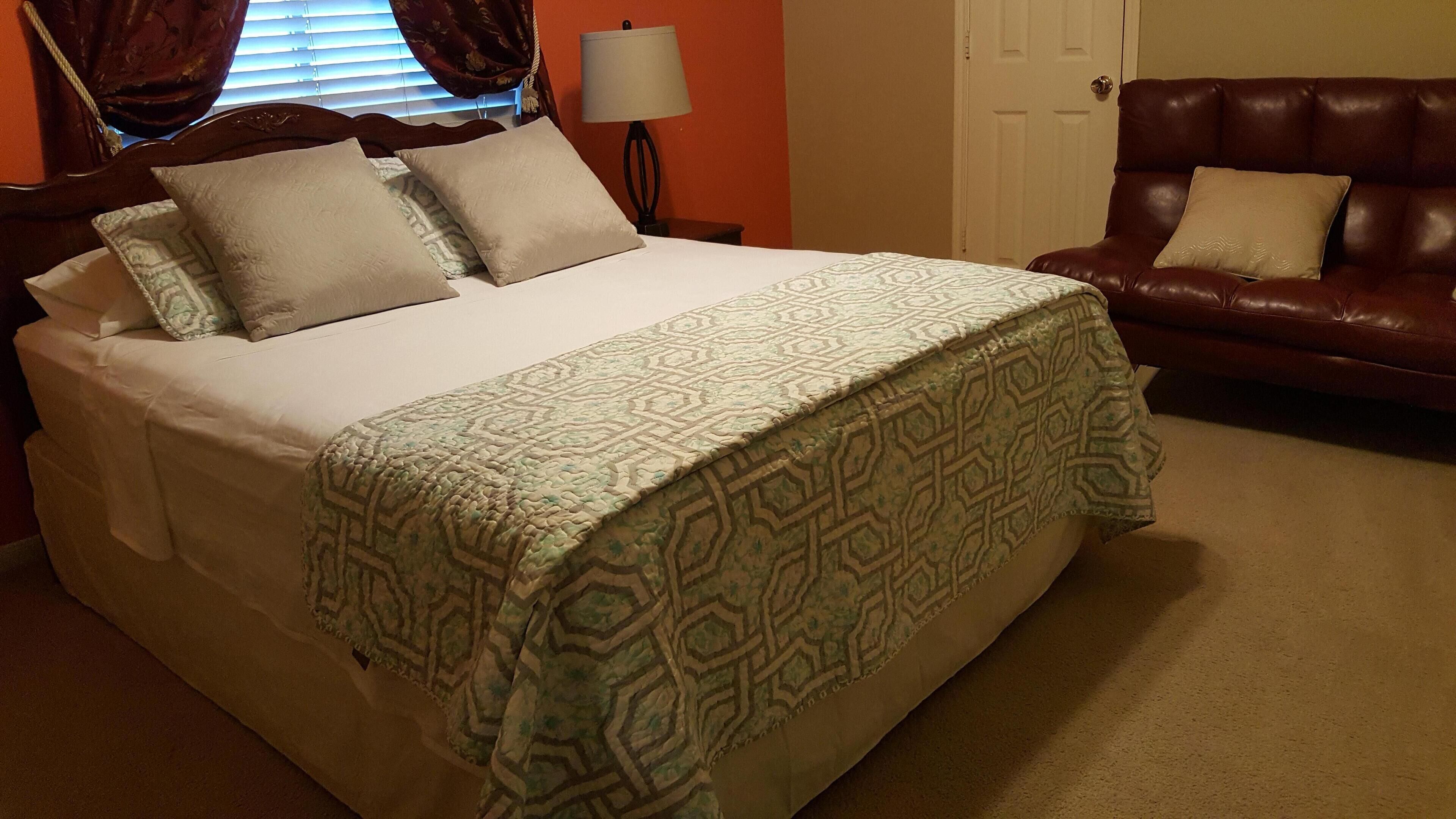A neatly made bed with a patterned bedspread, a brown leather sofa with cushions, and a lamp on a bedside table.