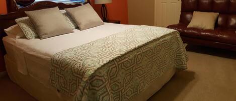 A neatly made bed with a patterned bedspread, a brown leather sofa with cushions, and a lamp on a bedside table.