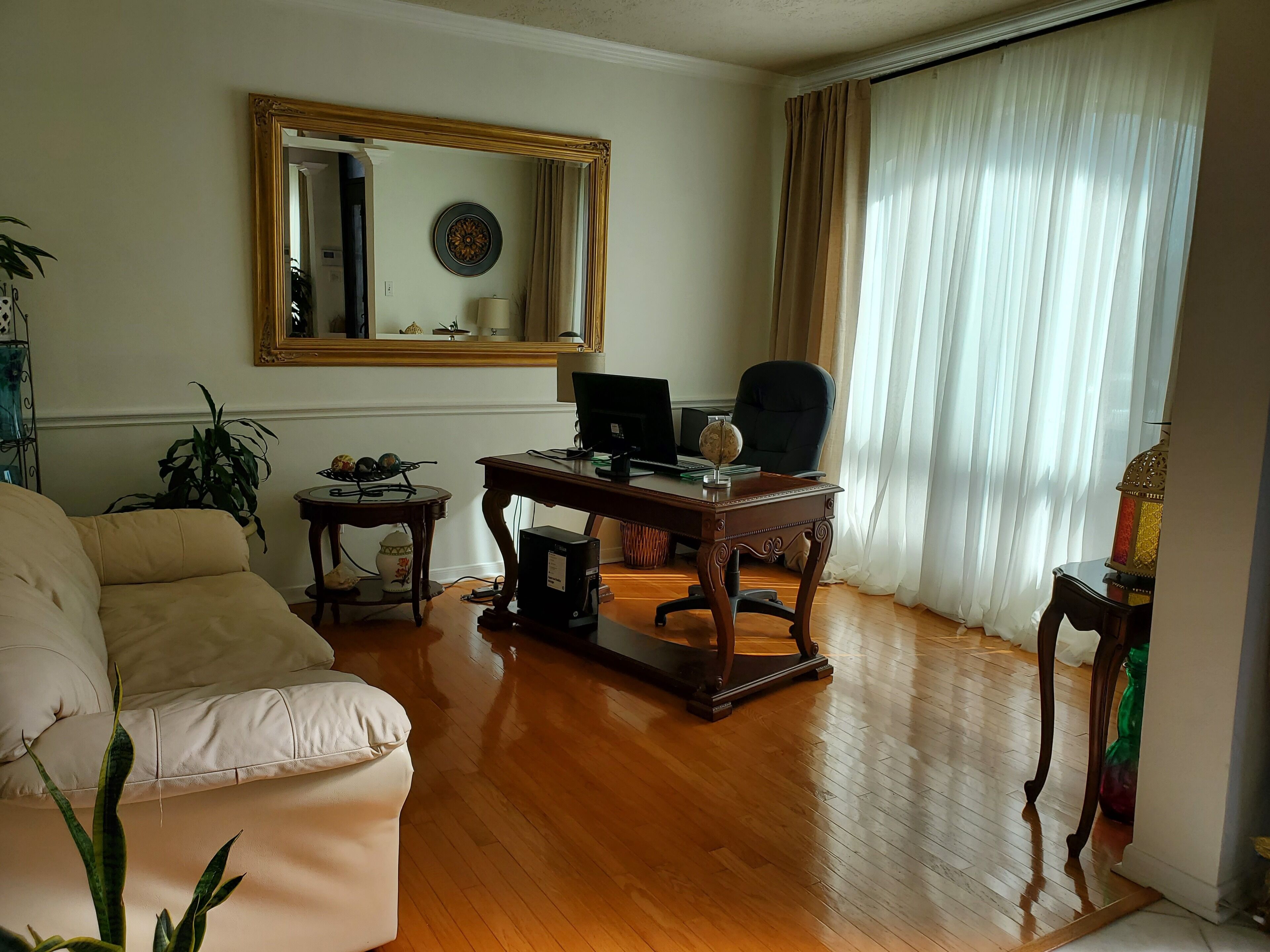 Een woonkamer met een bank, een bureau met een computer, een stoel en een groot raam met gordijnen.