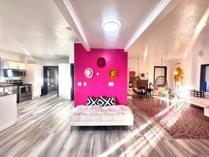 Un espacio habitable moderno con una pared de acento rosa vibrante, un sofá blanco y una cocina con electrodomésticos de acero inoxidable.