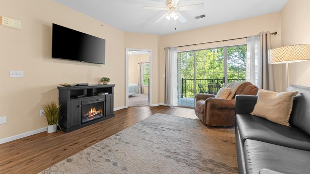 A living room with a flat-screen TV, a fireplace, a ceiling fan, and a view to a balcony. - Golf View Resort Condo | No Stairs + Pools & Perks (Branson)