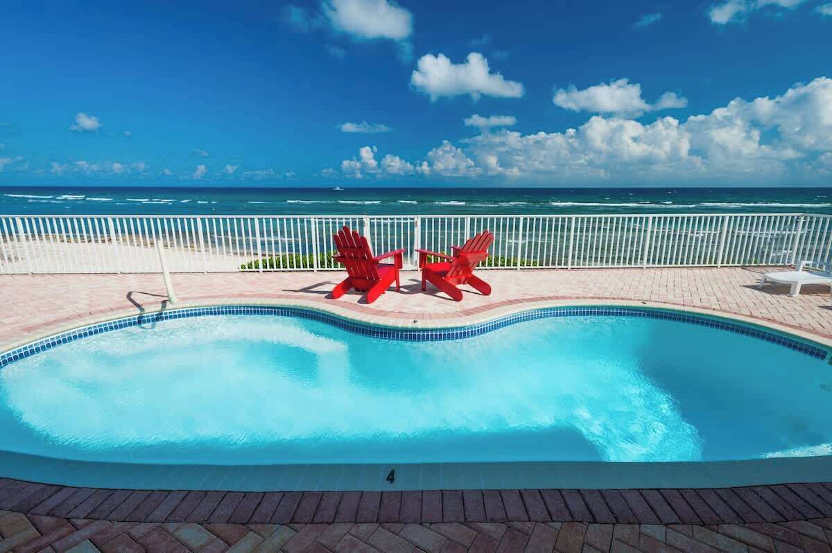 A swimming pool with two red Adirondack chairs facing the ocean.